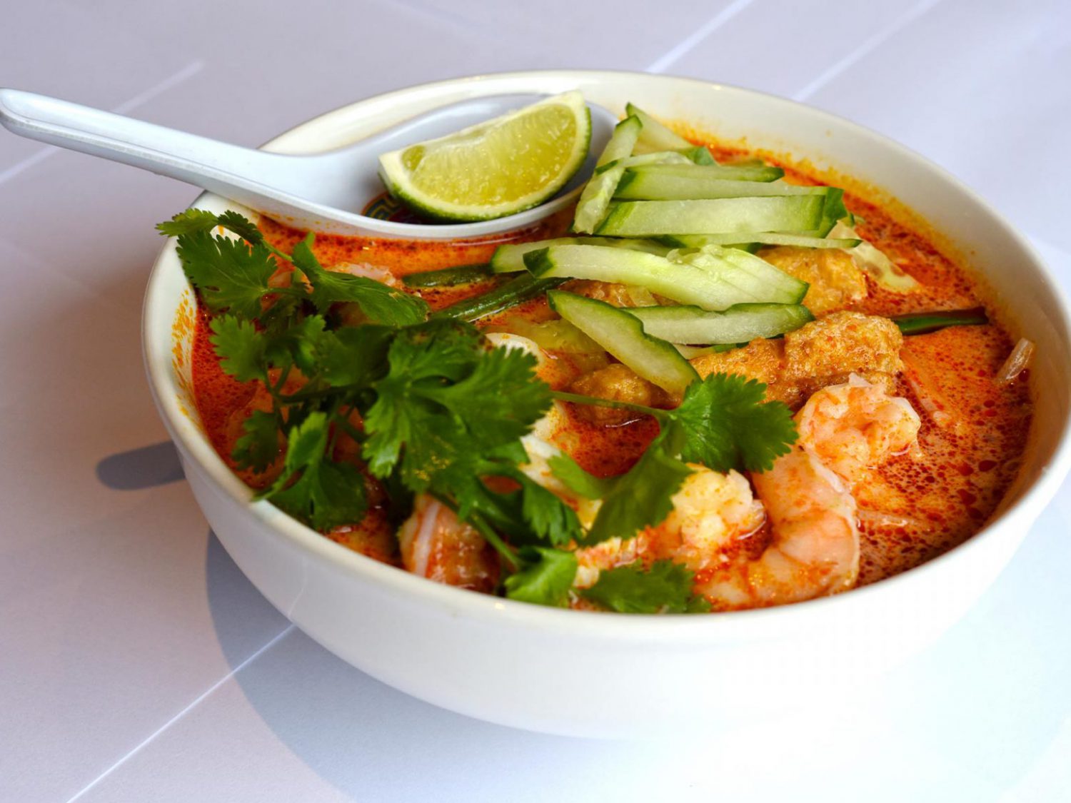 Soup bowl filled with prawns, coriander, cucumber slices and a slice of lime on top of the spoon