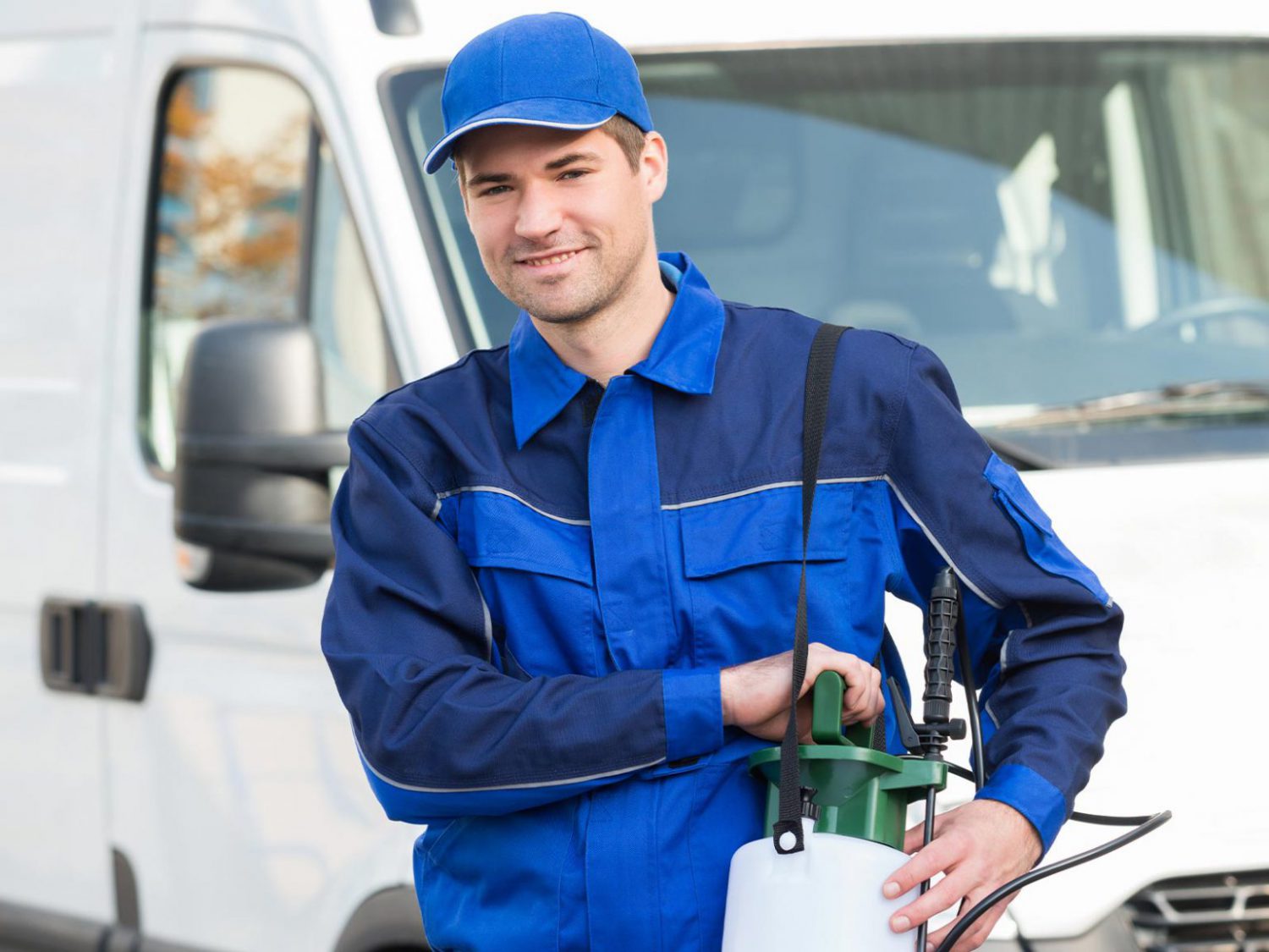 A man standing in frot of a white van, wearing a blue uniform and carrying some pest control equipment on his left shoulder