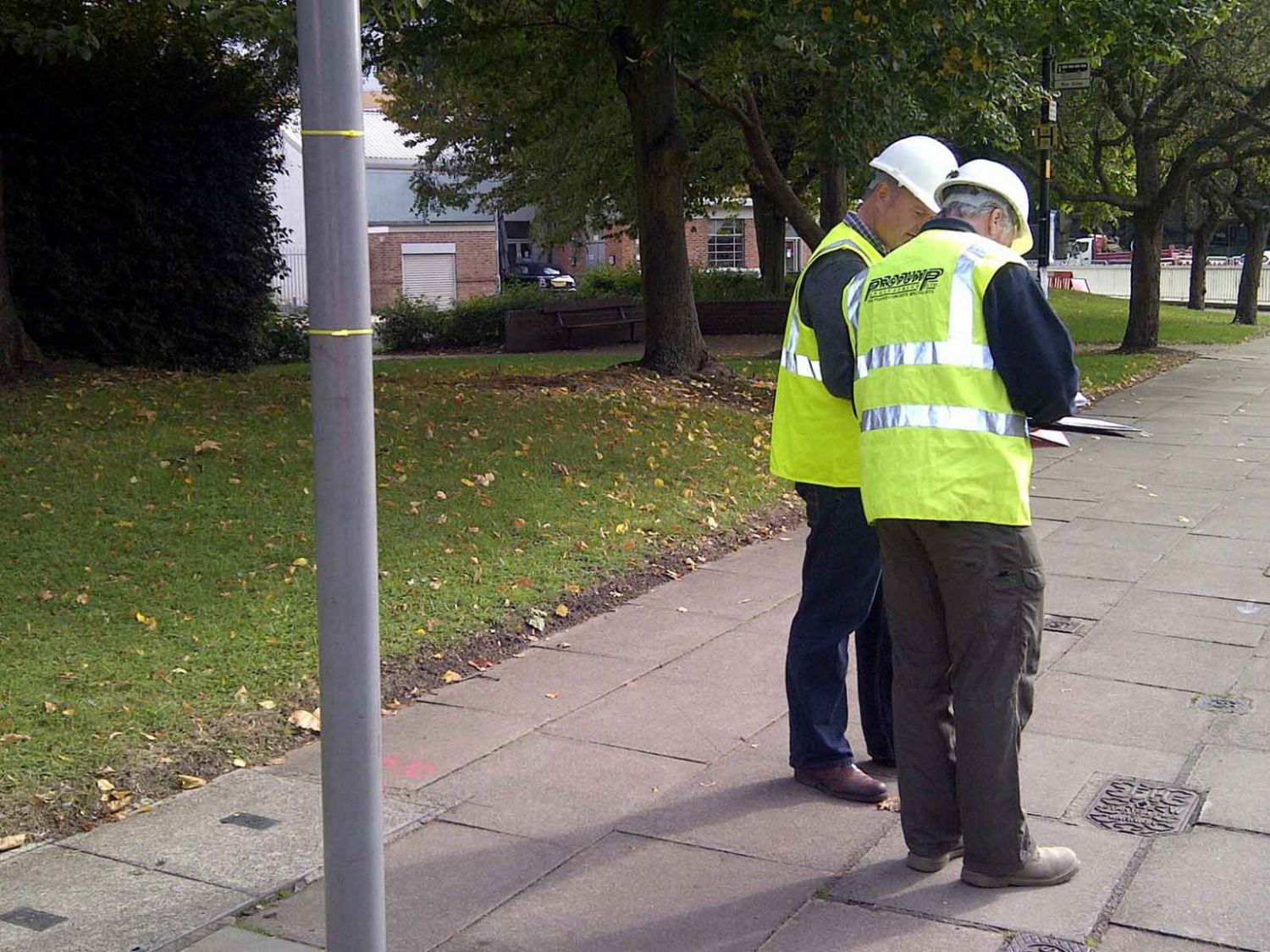 Two men wearing fluorescent yellow vests, wearing helmets whilst standing on the pavement checking some drawings