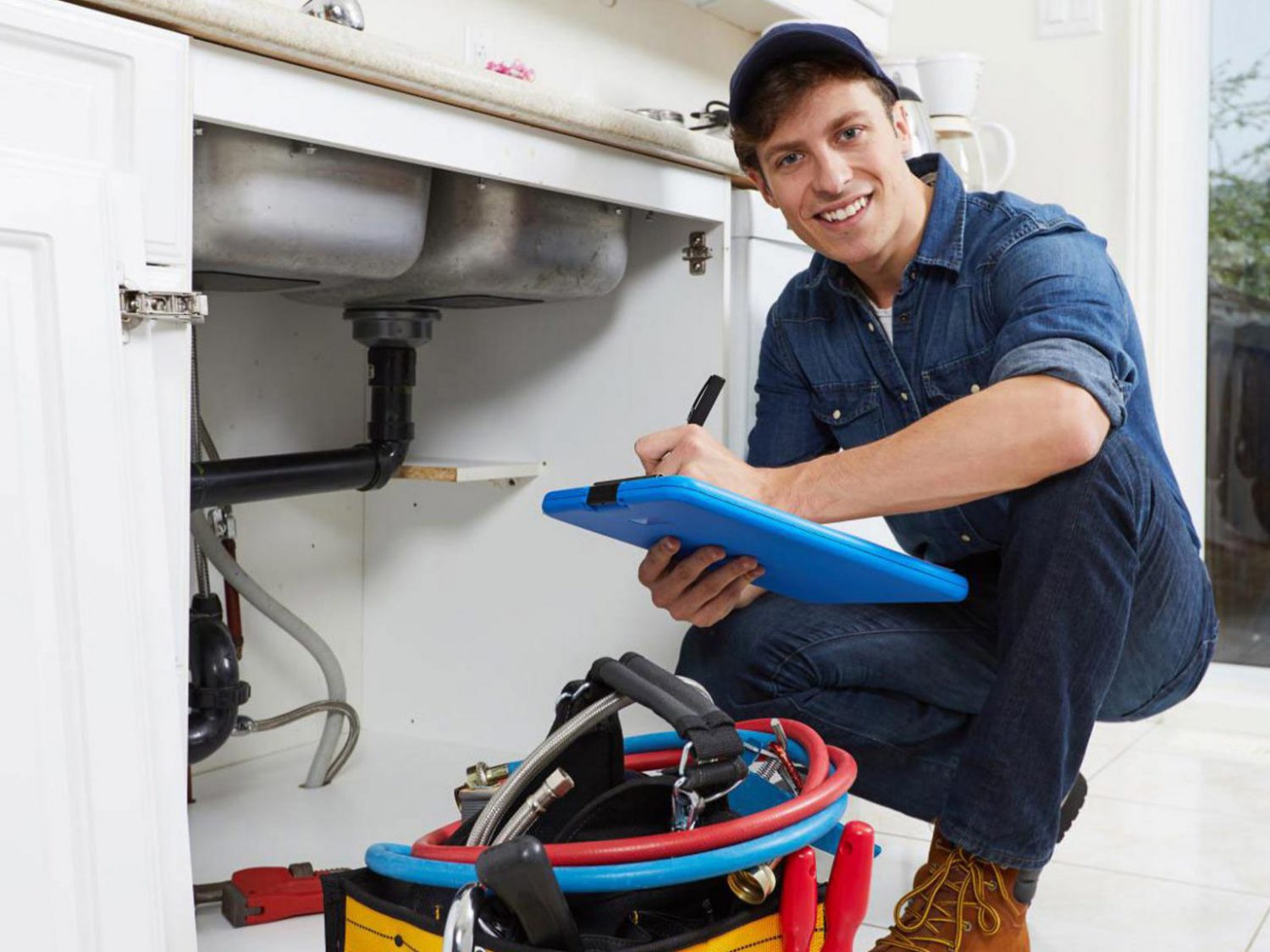 A smiley plumber in a squatting position, wearing denim overalls and taking notes on his notepad